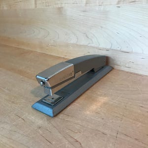 May include: A silver Bosstitch stapler with a gray base. The stapler is on a wooden surface.