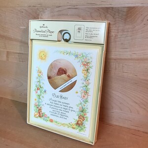 Puede incluir: Un marco de fotos blanco con un paspartú amarillo y un recorte para una foto. El marco tiene un diseño floral y el texto "Our Baby" con un poema sobre un bebé. El marco está hecho por Hallmark.