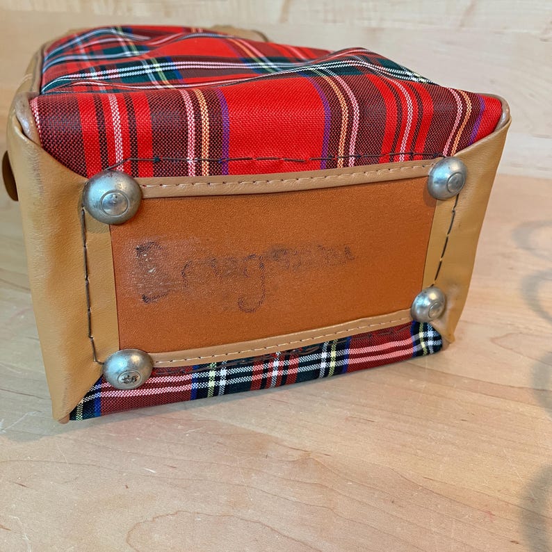 May include: A vintage travel bag with a red, green, and white tartan pattern. The bag features tan leather trim and metal feet. The base of the bag has a brown leather patch with faded text.