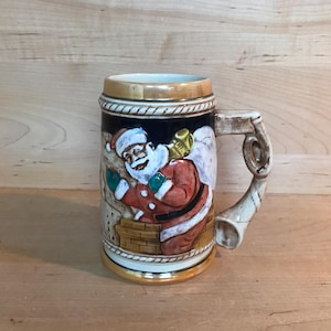 Puede incluir: Una jarra de cerveza navideña de cerámica con una representación detallada de Santa Claus bajando por una chimenea. La jarra tiene un asa marrón y un borde dorado.