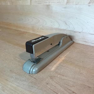 May include: A vintage Swingline stapler in a light gray color with a brown top. The stapler has a metal body and a plastic top with the Swingline logo in black lettering. The stapler is designed to hold standard size staples.
