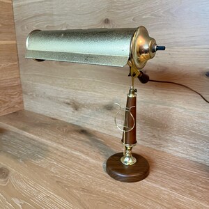 May include: A vintage desk lamp with a gold-toned shade and accents. The lamp features a dark wood base and stem, with a decorative gold-colored lyre-shaped detail. The lamp is designed to be adjustable and has a black on/off switch.