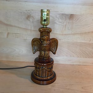 Könnte beinhalten: Eine braune Keramik-Tischlampe mit Adler-Design. Die Lampe hat eine goldfarbene Fassung und ein braunes Kabel.