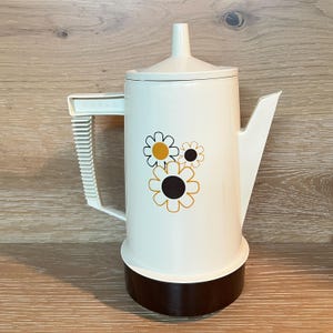 May include: Cream-coloured vintage coffee pot with a brown base and handle. The pot features a floral design with yellow and black accents. The pot has a lid and a spout for pouring.