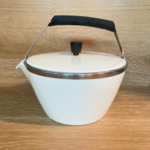 May include: A white teapot with a smooth, tapered body and a silver-toned metal rim. It features a black lid knob and a black handle attached to a silver metal frame that arches over the top. The teapot has a small, integrated pouring spout.