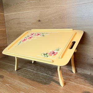May include: A light yellow breakfast tray with folding legs and integrated side handles. The surface features a white grid pattern accented with pink floral motifs in two corners. The tray includes a raised edge border and a front ledge support.