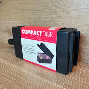 May include: A black plastic compact disk tote with a red paper sleeve featuring the text COMPACT DISK TOTE. The sleeve highlights a durable clam-shell design, fits under or between car seats, and organizes and protects 12 compact discs. Made in USA.