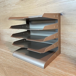 Könnte beinhalten: Ein metallischer, bronzefarbener Schreibtisch-Organizer mit fünf horizontalen, gestuften Ablagen zur Dokumentenaufbewahrung. Das Gerät verfügt über eine vertikale Rückwand mit seitlichen Stützen, die jedes Fach in einer festen, schrägen Position halten.