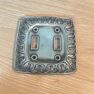 May include: A silver-toned, ornate light switch plate with a decorative floral border. The plate has two toggle switch openings and four screw holes. The plate is made of metal and has a vintage aesthetic.