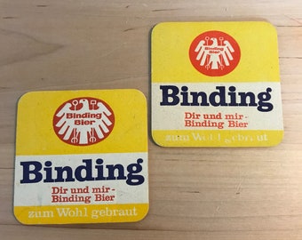 以下が含まれることがあります： 赤い白の鷲のロゴと青い「Binding Bier」の文字が書かれた、黄色の正方形のビールコースター2枚。コースターには「Dir und mir - Binding Bier zum Wohl gebraut」の文字も印刷されています。