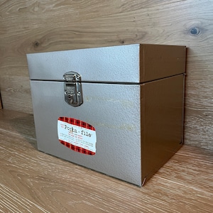 Puede incluir: Una caja Porta-file de metal plateado con cierre de pestillo. La caja tiene una etiqueta con el texto "Porta-file" y detalles de cuadros rojos. Diseñada para uso doméstico o de oficina.