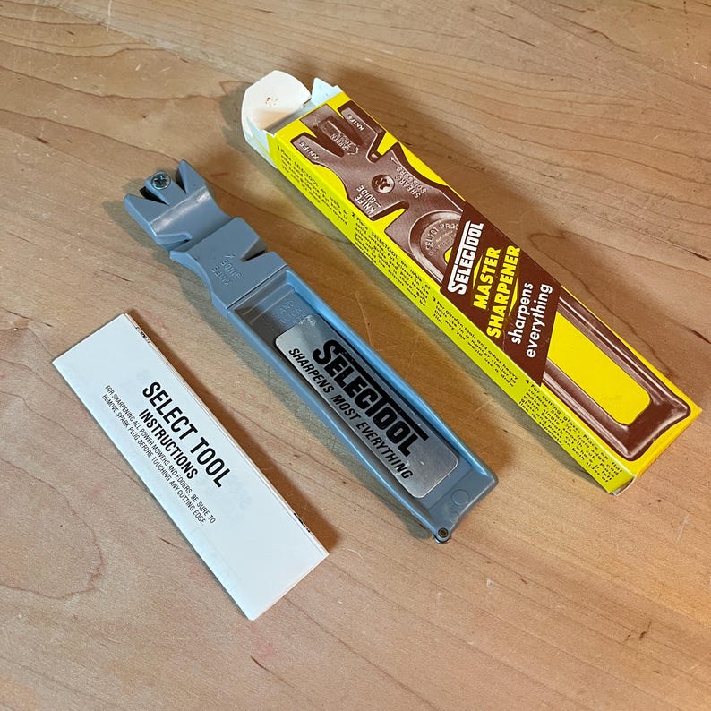 May include: A gray Select Tool knife sharpener with the text "SHARPENS MOST EVERYTHING". The packaging includes a yellow and brown box with "SELECTOOL MASTER SHARPENER" printed on it, and a white instruction sheet.