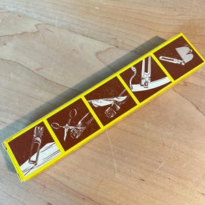 May include: A rectangular yellow and brown box with illustrations of tools. The box has a yellow border and brown panels with white line drawings. The box is likely for a craft or sewing kit, with images of scissors and other tools.