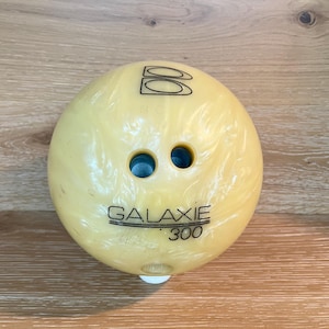 May include: A yellow bowling ball with a swirling pattern. The ball has three finger holes and the text "GALAXIE 300". The top of the ball has the letter "B" and two overlapping rectangles.