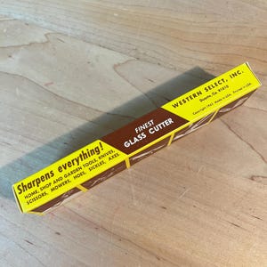 May include: A yellow and brown box for a glass cutter. The box has text that reads "Finest Glass Cutter" and "Sharpens everything!" It also lists various tools. The box is from Western Select, Inc.