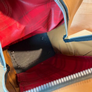 May include: An open brown leather bag with a red interior lining. The bag has a black base and a silver zipper. The interior is visible, showing the stitching and construction of the bag.