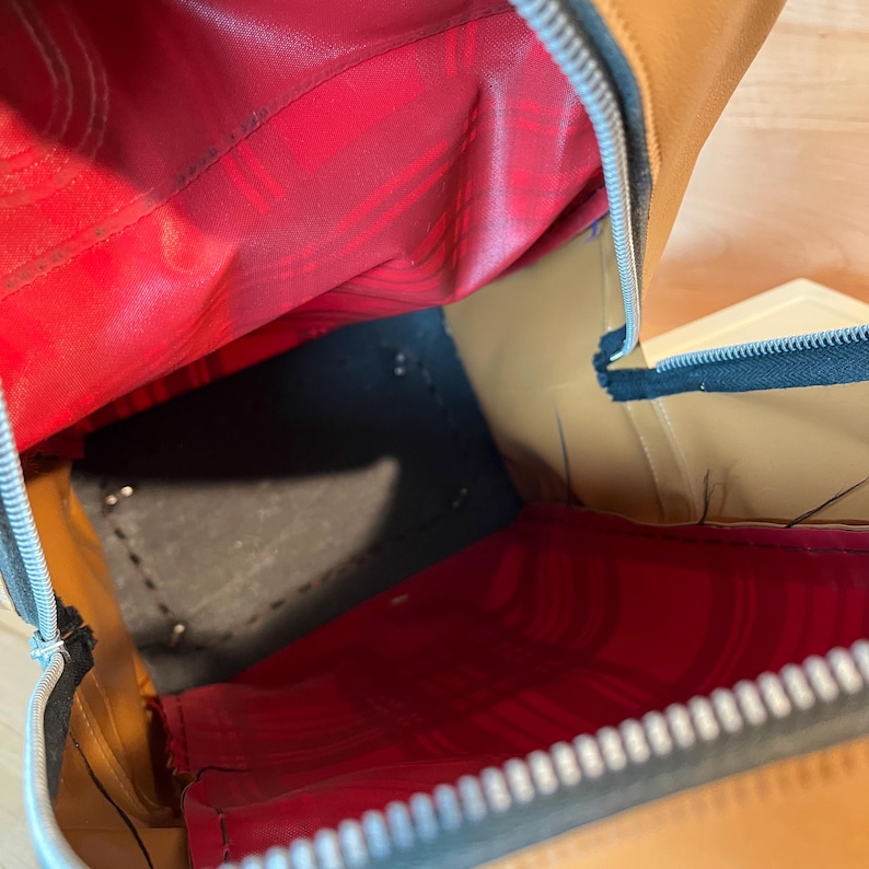 May include: An open brown leather bag with a red interior lining. The bag has a black base and a silver zipper. The interior is visible, showing the stitching and construction of the bag.