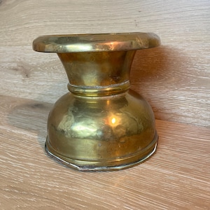 Puede incluir: Un portavelas de latón vintage con una parte superior ancha y plana y una base bulbosa. El portavelas tiene un tono dorado cálido y muestra signos de edad y uso. El diseño es simple pero elegante, adecuado para varios estilos de decoración.