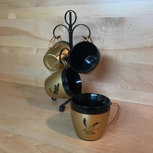 May include: Set of four gold and black coffee mugs with a bird design. The mugs are hanging on a black metal stand.