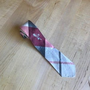 May include: A gray and pink patterned tie with a burgundy patch featuring a quill and scroll design. The patch has the text "University of Notre Dame" on it.