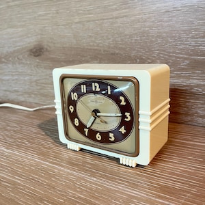 Puede incluir: Despertador vintage color crema con esfera marrón y beige. El reloj tiene forma cuadrada con bordes redondeados y un borde dorado. La esfera del reloj presenta números y manecillas negros, con las palabras "General Electric" impresas.