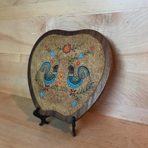 May include: Heart-shaped trivet with a cork centre and a wooden frame. The trivet features a colourful illustration of two roosters, flowers, and foliage. The roosters have blue, yellow, and orange details. The trivet is displayed on a black stand.