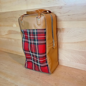 May include: A vintage travel bag with a tan leather exterior and a red plaid fabric panel. The bag has a top handle, a full-length zip, and metal hardware. The bag is rectangular and stands upright.