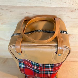 May include: Tan leather travel bag with a red, green, and white plaid pattern on the bottom. The bag has a zip closure and two brown handles. The bag is on a wooden surface.
