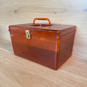 May include: A rectangular, translucent amber-colored plastic storage box with a hinged lid and a top handle. The box features a textured, woven-like pattern on the exterior surfaces and is secured with a central gold-toned metal latch.