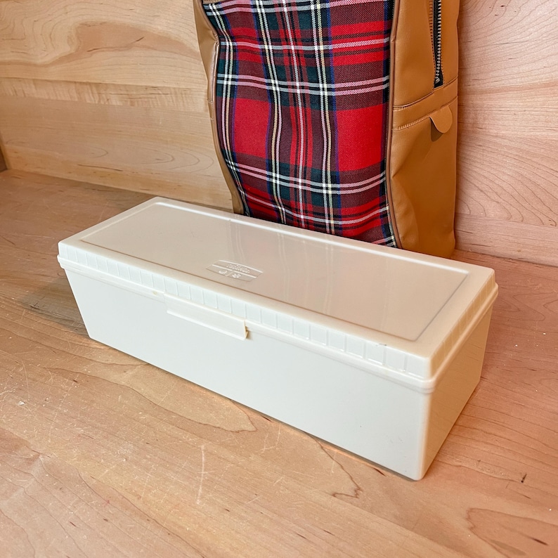 May include: An off-white plastic storage box with a lid, rectangular in shape. The box sits on a wooden surface. Behind it is a brown and plaid tote bag. The tote bag has a zip and a leather-like texture.