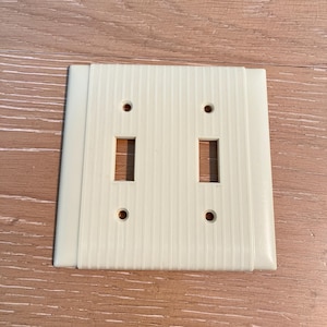 以下が含まれることがあります： オフホワイトのダブルスイッチカバー。中央にリブ模様のデザイン。2つの長方形のスイッチ開口部と4つのネジ穴があります。エッジは滑らかで丸みを帯びています。