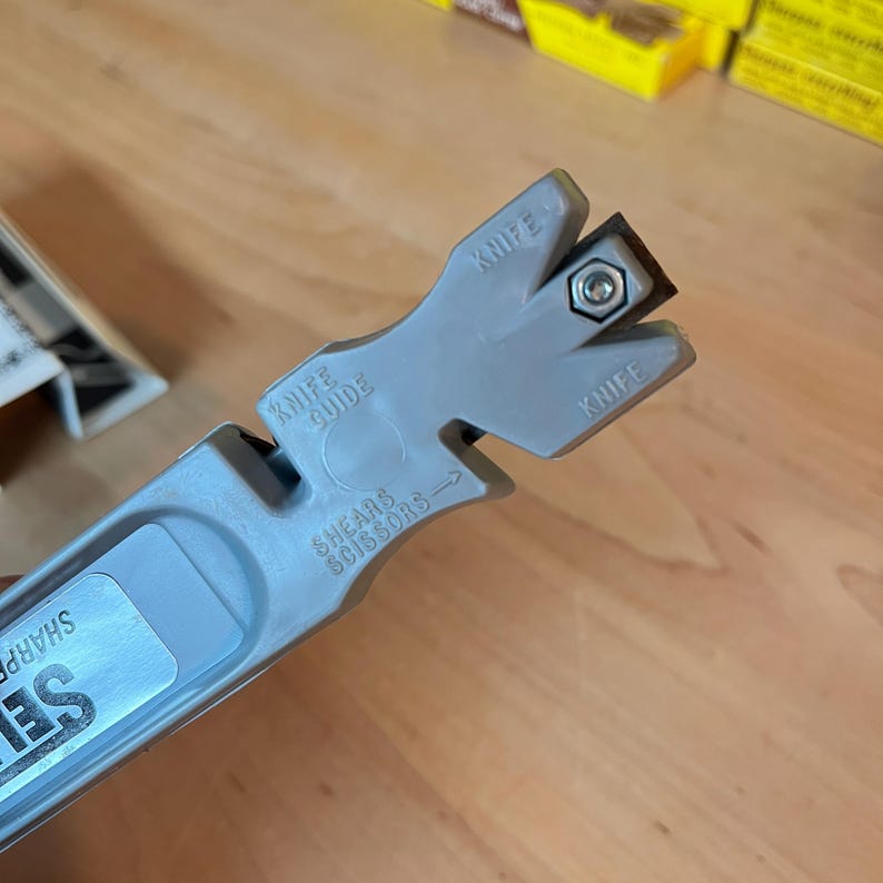 May include: A gray knife and scissors sharpener with the text "Knife Guide" and "Shears Scissors". The sharpener has a slot for the knife and a small sharpening stone. The tool is designed to sharpen knives and scissors.