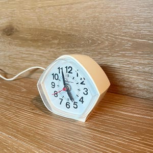 May include: A vintage peach-coloured alarm clock with a white face and black numbers. The clock has a clear plastic cover and a red second hand. The clock is on a wooden surface.