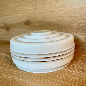 May include: A white, circular light fixture with a layered design. The fixture has gold-colored bands around the base and a clear, circular top. The fixture is sitting on a wooden surface.