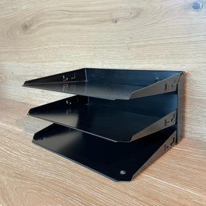May include: A black metal desk organiser featuring three stacked horizontal trays. The unit has a solid vertical back panel with integrated slots for adjusting the tray positions, providing a tiered storage solution for documents and office supplies.
