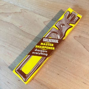 May include: A yellow and brown Selectool Master Sharpener box. The box features the text "Selectool Master Sharpener sharpens everything" and a diagram of a knife guide. The box is angled on a wooden surface.