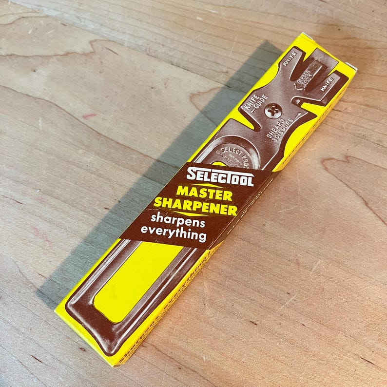 May include: A yellow and brown Selectool Master Sharpener box. The box features the text "Selectool Master Sharpener sharpens everything" and a diagram of a knife guide. The box is angled on a wooden surface.