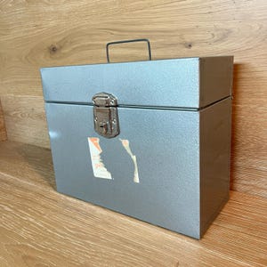 May include: A rectangular silver metal storage box with a hinged lid and a black wire handle. The front features a central silver metal latch with a keyhole and a partially torn white paper label with faint orange text.