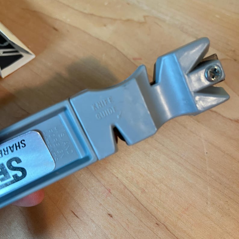 May include: A gray knife sharpener with the text "KNIFE GUIDE" printed on the side. The sharpener has a plastic body with a metal screw and a slot for sharpening blades. A sticker with the word "SHARP" is partially visible.