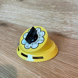 May include: A bright yellow kitchen timer with a slanted, circular face shaped like a flower. It features a black central knob and a white dial marked with numbers 15, 30, and 45, along with the text "time mark" and "turn past" printed in black.