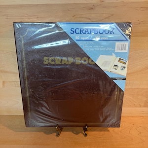 Puede incluir: Un álbum de recortes marrón con detalles dorados y el texto "Scrapbook 50 sheets / 100 pages" en la portada. El álbum está envuelto en plástico y tiene un código de barras en el lateral.