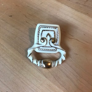 Puede incluir: Tirador de armario de cerámica blanca con un diseño adornado de color dorado. El tirador tiene una forma cuadrada con un asa redondeada.