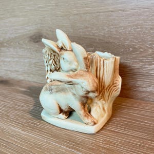 May include: A ceramic planter featuring two rabbits and a tree stump design. The rabbits are light brown and cream-colored, with detailed features. The tree stump is a light brown color. The planter is a decorative item.