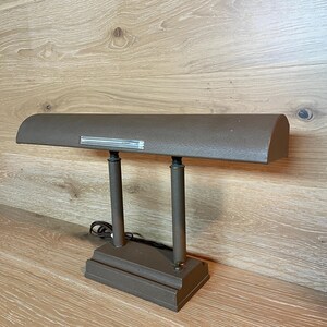 May include: A brown desk lamp with a rectangular base and two vertical supports. The lamp has a curved shade with a silver accent. The lamp is made of metal and has a power cord.