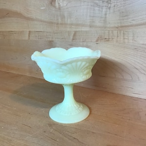 May include: A small, pale yellow glass pedestal bowl with a scalloped rim and a decorative floral design. The bowl is on a wooden surface.