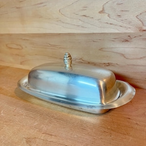 May include: Silver butter dish with a lid and a decorative pinecone-shaped handle. The dish has an oval shape and sits on a matching base. The butter dish is made of a shiny, reflective metal.
