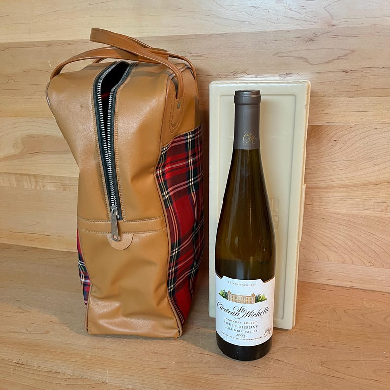 May include: A tan leather wine carrier with a red and black tartan accent, a bottle of Chateau Ste. Michelle Sweet Riesling wine, and a white wine case. The wine bottle has a white label.