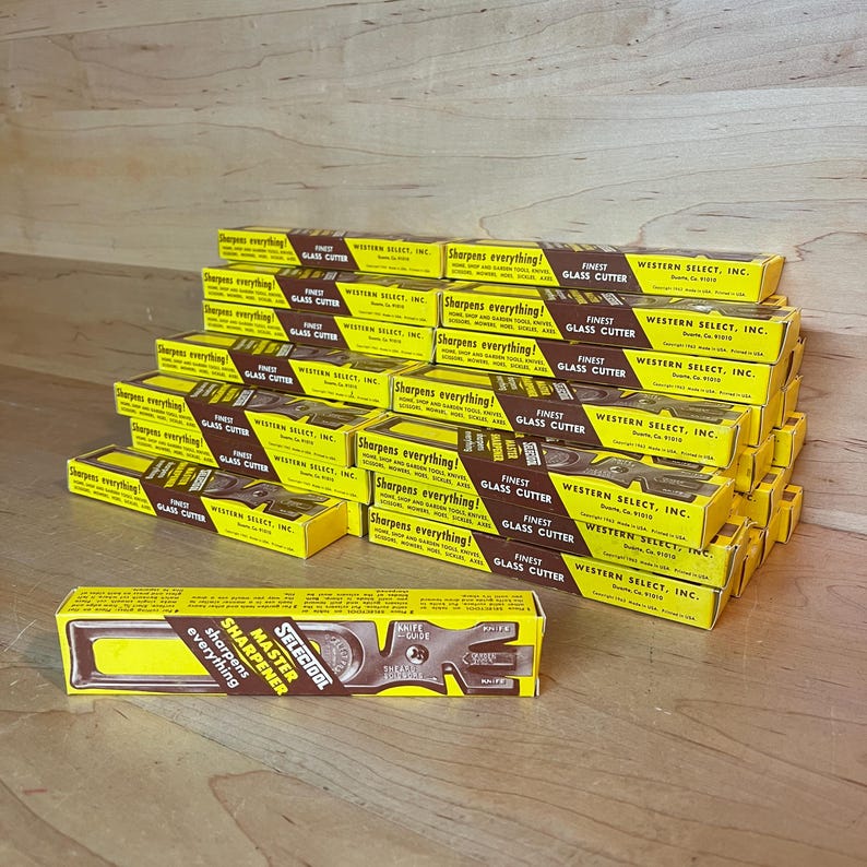 May include: A collection of yellow and brown boxes of glass cutters and sharpeners. The boxes are stacked, with text that reads "Sharpens everything!" and "Finest Glass Cutter". The brand name "Western Select, Inc." is also visible.