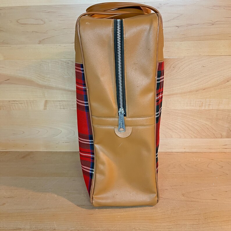 May include: A vintage tan travel bag with a black zip and a red, green, and white plaid pattern on the sides. The bag has a handle and is made of a leather-like material.