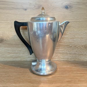 Könnte beinhalten: Eine silberfarbene Vintage-Kaffeekanne mit schwarzem Griff und einem Deckelknauf aus klarem Glas. Die Kanne hat einen Ausguss zum Ausgießen und einen Sockel mit einem elektrischen Stecker. Die Kaffeekanne ist aus Metall und hat ein klassisches Design.